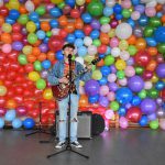 Młody mężczyzna z gitarą śpiewający do mikrofonu, w tle ściana z kolorowych balonów
