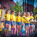 Występ artystyczny. 8 dziewczynek w żółtych koszulkach śpiewa do mikrofonów