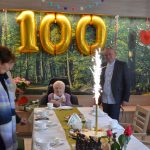 Na zdjęciu stulatka Szarlotta Moll z bliskimi, na stole tort z zapalonym fajerwerkiem, w tle balony ułożone w liczbę 100