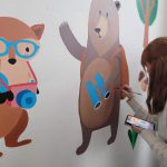 Dziewczyna maluje na ścianie niedźwiedzia. Obok jest już namalowany lisek w niebieskich okularach.