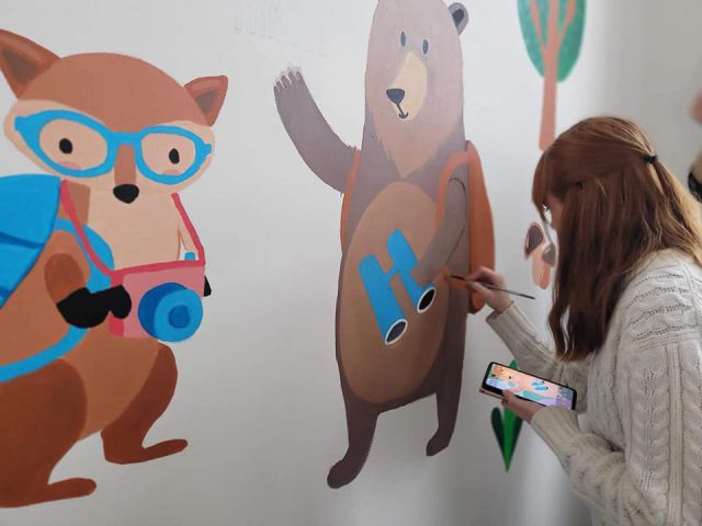 Dziewczyna maluje na ścianie niedźwiedzia. Obok jest już namalowany lisek w niebieskich okularach.