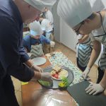 Uczniowie w kucharskich czapkach wraz z Remigiuszem Rączką przygotowują kanapki
