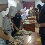 Uczniowie w kucharskich czapkach przygotowują kanapki. Remigiusz Rączka ogląda ich pracę