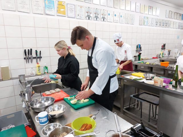 Uczniowie w fartuchach kucharskich szykują jedzenie w pracowni gastronomicznej.