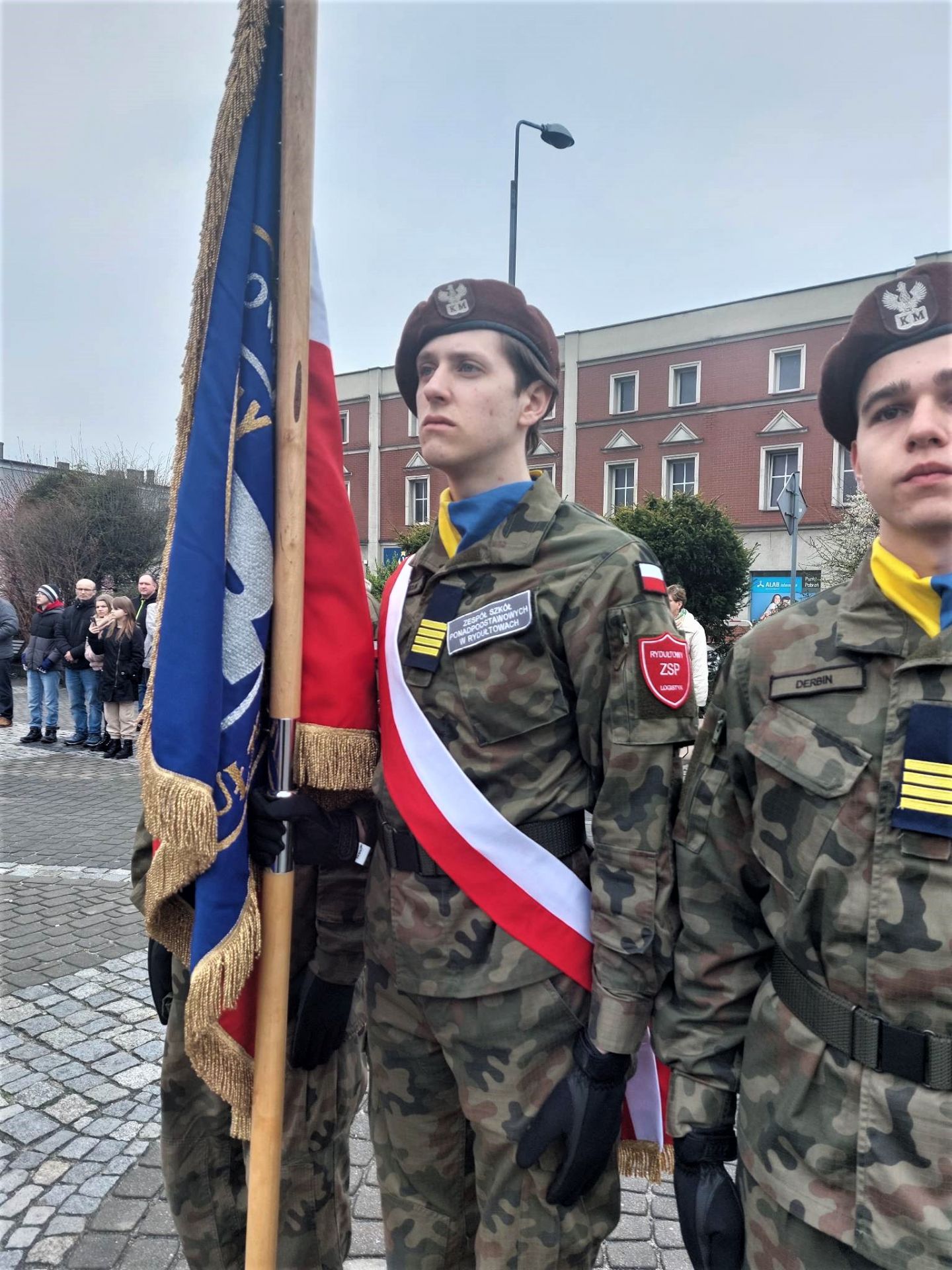 Na zdjęciu poczet sztandarowy szkoły, składający się z 3 młodych osób ubranych w mundury wojskowe, trzymających sztandar szkoły