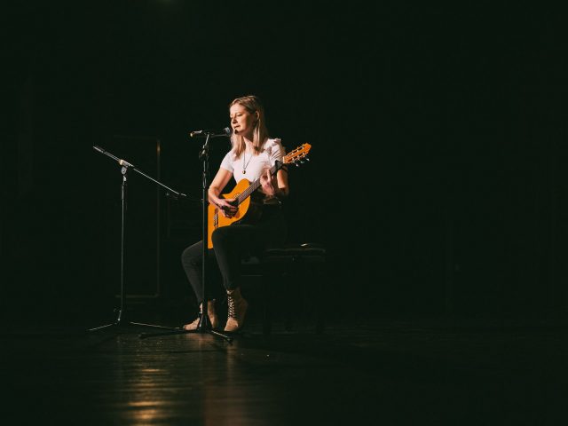 Uczennica ubrana w białą koszulkę siedzi na krześle, gra na gitarze, jest na scenie, na scenie panuje półmrok.