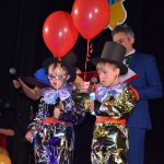 Dwóch chłopców stoi na scenie, w rękach trzymają czerwone balony.