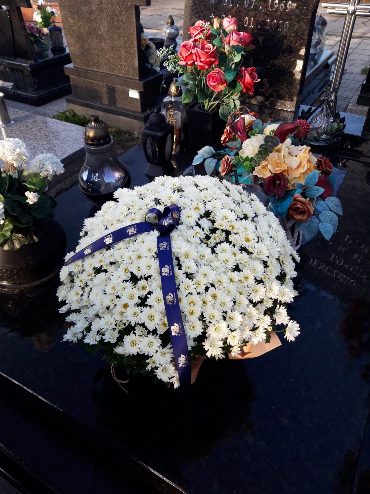 Doniczka białych chryzantem przepasana wstęgą z napisem 25 lat Powiatu Wodzisławskiego ustawiona na pomniku nagrobnym