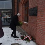 Złożenie kwiatów w miejscach pamięci Ofiar Marszu Śmierci z Auschwitz Birkenau do Wodzisławia Śl. Na zdjęciu 2 postacie ludzkie przed tablicą pamiątkową na dworcu kolejowym. Przewodniczący rady Adam Krzyżak składa wiązankę pod miejscem pamięci