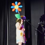 Dziewczyna stoi na scenie i śpiewa do mikrofonu. W rękach trzyma dużego kwiatka