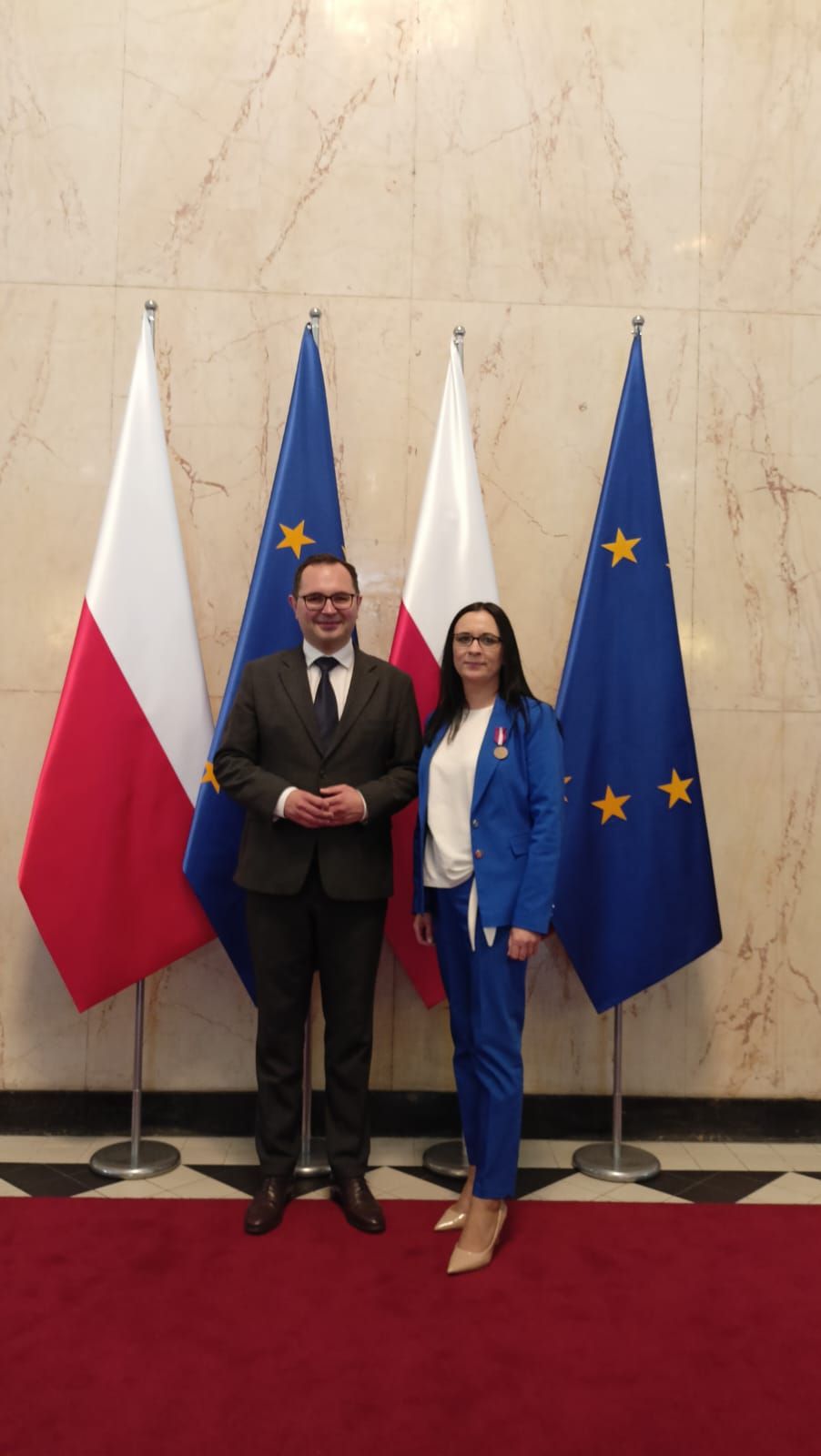 Kobieta i mężczyzna stojący na tle flag Polski i Unii Europejskiej. Kobieta w klapie marynarki ma wpięty medal