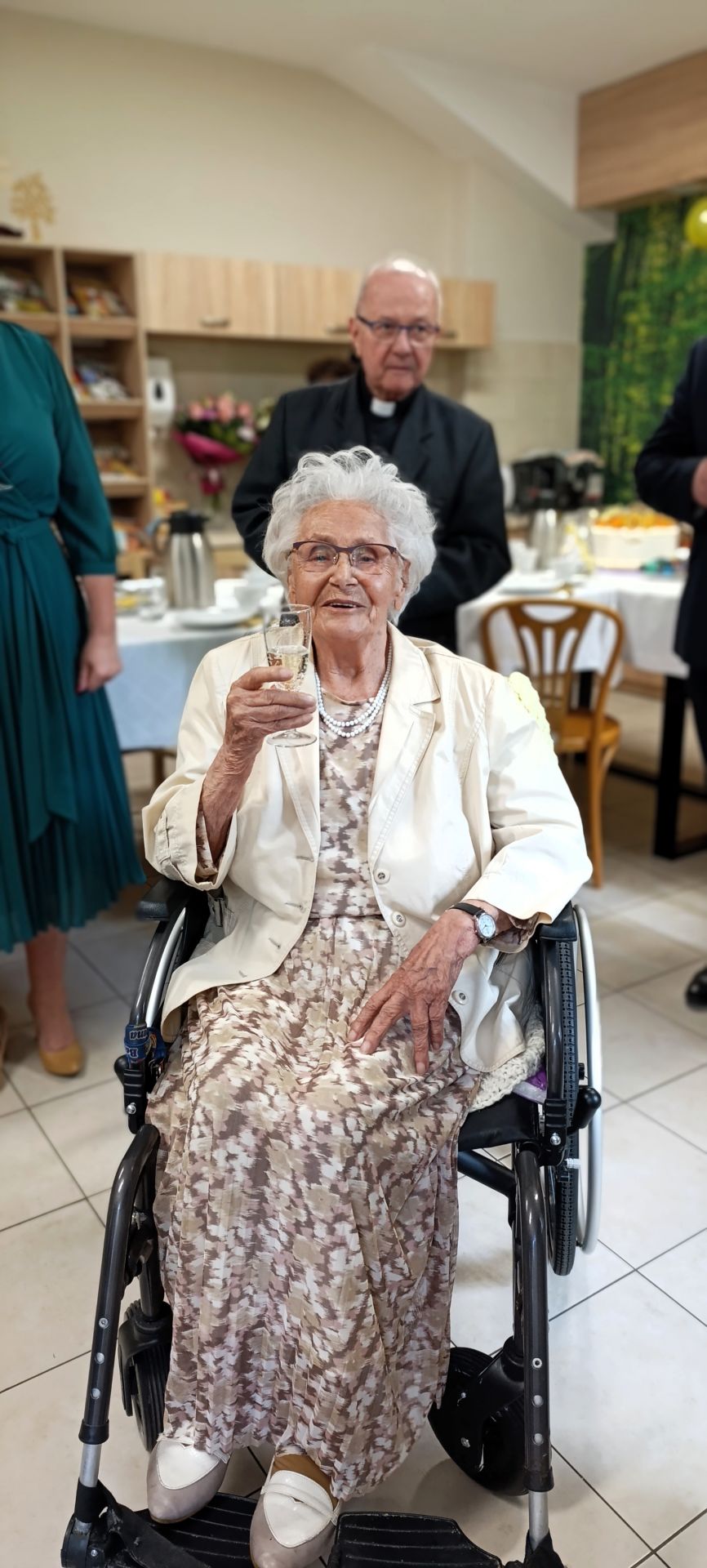 Jubilatka wznosi toast, siedzi na wózku inwalidzkim, ubrana w jasną sukienkę i białą marynarkę