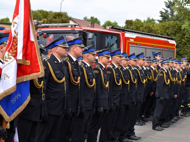 Strażacy w galowych mundurach stoją w szeregu. Jako pierwszy stoi poczet sztandarowy