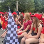Grupa młodych dziewczyn przebranych w czerwone stroje z logo ferrari i trzymających flagę zapowiadającą wyścig formuły 1