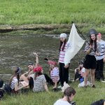 Grupa młodzieży odpoczywającej nad rzeką. Jedna osoba trzyma w ręce flagę. Młodzież jest ubrana w pirackie stroje